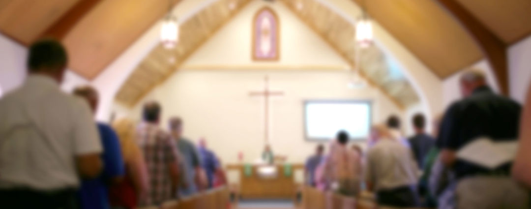 A blurred photo of the inside of a church sanctuary that is filled with people in the pews, and the pastor stands under a large cross at the altar.